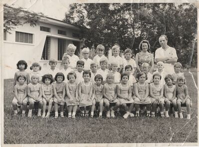 Tengah Primary school photo, of year 1 - 1964
I am in front of the gentleman who I think was the head teacher.
 I think the photo was taken in the grounds between my class and the school hall, my class room is in the background, I expect the hall to be to the right out of camera.
You will see on my neck a white line. That is real, it is not a crease in the picture, I had had throat surgery a couple of months before flying out to Singapore to remove a growth in the larynx  glottis as I had stopped talking.  
Unrelated there is also  white marks  above and below my right eye, that is just two of fifty others from a dog attack about a year before, I still looked a mess and was still getting patched up, and spend a fair amount of time at the forces hospital having things fixed. There I didn't like the air conditioned ward it was too cold and insisted on being put in the native patents area. My Mon had an argument with them to alwo me to change to the natives area and also later helped me get the native food rather than the english stuff.  The one hospital meal I remember was leaks, seriously over cooked but the leeks were good compared to everything else. At hospital I looked forward for my familys chinese Amah  Cheng to visit, because Cheng usually brought some of her chinese cooking in a little basket.
Shortly before the school picture was taken I remember a fire drill where the head teacher brought a smoking litter bin into the class.
I remember the small milk bottles and the strawberry and chocolate milk.
Later I changed class which was in the main school just on the other side to my first class. The milk was delivered in the corridor between our two classes. I sort of got into trouble when I went to my old class milk box and took a strawbury milk, and the teacher in the photo explained very nicely that my class does not  get flavored milk, but I did get to keep the milk that once. 
I think we also got orange juice in triangular pyramid paper cartons, but I may be confusing with when my dad was at  USAF sorry RAF Fylingdales.
I also remember a couple of lessons in one we talked about what England was like as it was winter there, cold, lots and lots of snow, very dark, stormy, and big waves, we then did a session painting. Another we talked about Astronauts and rockets. 
The main school was three sides of a square, with a covered walkway (cloister), the center section had an entrance in the middle that went to a Huge hall that had two rows of fans in the ceiling which moved and a row big fans in the center. In the cloister to the right of the hall entrance was a huge red coke machine.
I also remember the playing fields and a sports day were I failed horribly at the  egg and spoon it was the first time i saw a concrete egg, then also the sack hopping failing with a full face splash, and finally the skipping I just kept the rope behind me and ran and won. I won a dinky toy bus which was presented  in the hall. It didn't feel good as I felt I had cheated with the skipping, but everyone seamed so nice and happy with the race.
We usually went to school and back in a bus  and the older kids sang Beatles songs, particularly "she loves you, yeh yeh yeh". However there was one day when the Army picked us up at school to take us home in lorries with propellers on the back, I remember being lifted up by a solder and handed bodily to another. who put me in behind a canvas cover.
There only one name from school I remember, that is Marvin and I think he is the one with the glasses in the photo. He lived at the end of my street in another bungalow like mine. I went to his birthday party and particularly remember the choice of red or green jelly and ice cream. 

Ironically when I returned to the yorkshire, it was in the depths of the worst winter in years, to go anywere I was wrapped up in about 10 layers of cothing I was freezing.

Keywords: Tengah;Primary School;1964;Neil Cooke